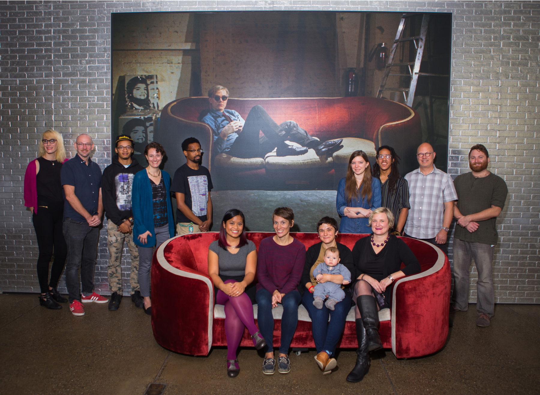 A group of staff members pose for a photo on and around a red couch. On the wall behind the couch is a photo of Andy Warhol lounging on a similar couch. One of the people on the couch is holding an infant.