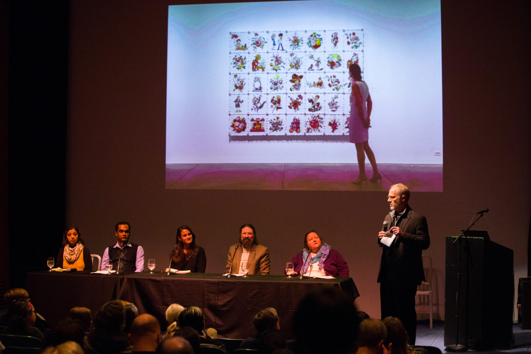 Five panelists are sitting at a table on the left side of a stage. They look towards a man who is standing and speaking into a microphone. Projected behind them is a photo of a woman looking at a large artwork comprised of a 5 by 7 grid of smaller illustrations.
