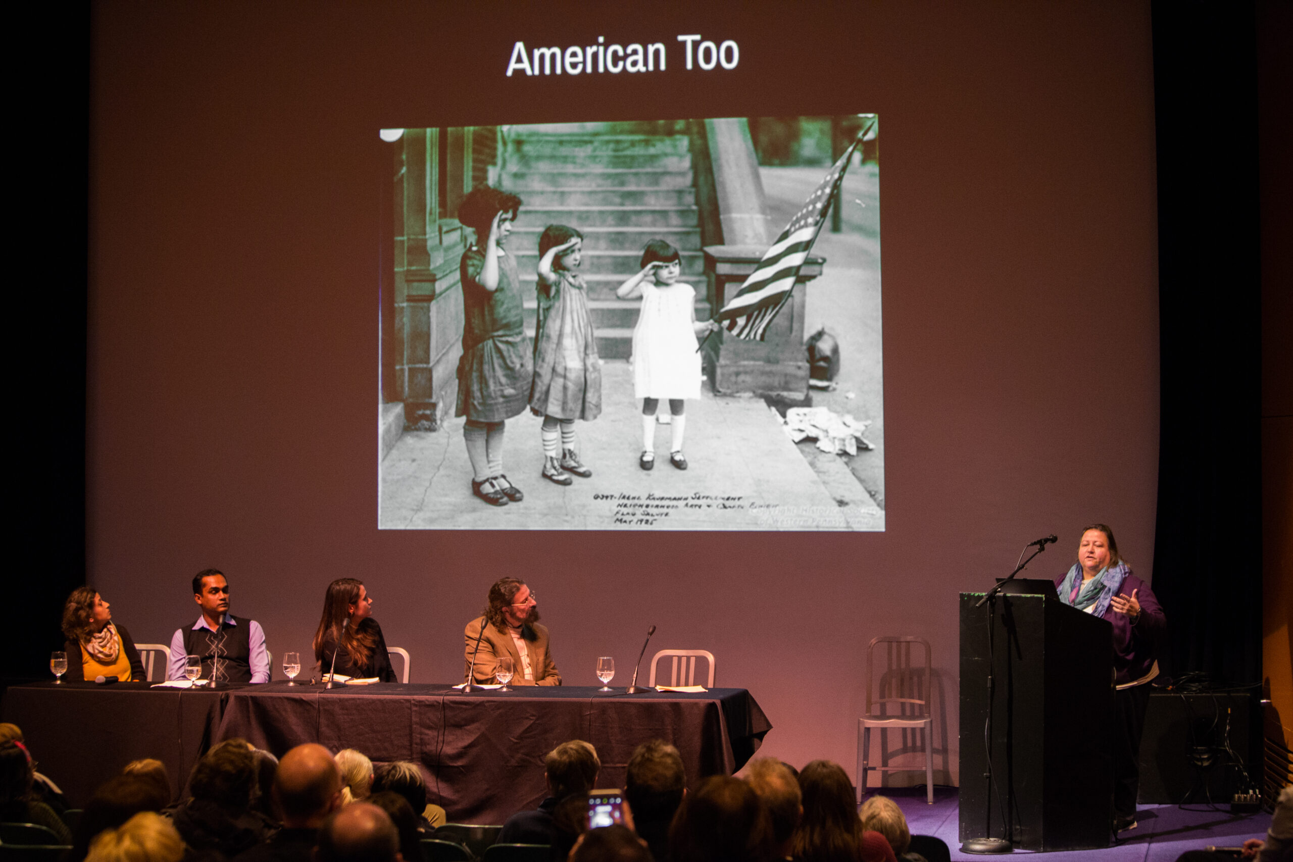 Four panelists sit at a table on the left-side of a stage. They are looking towards a woman speaking behind a lectern to the right. Behind them is projected a black and white image of three girls saluting an American flag. Above the image is the text American Too.