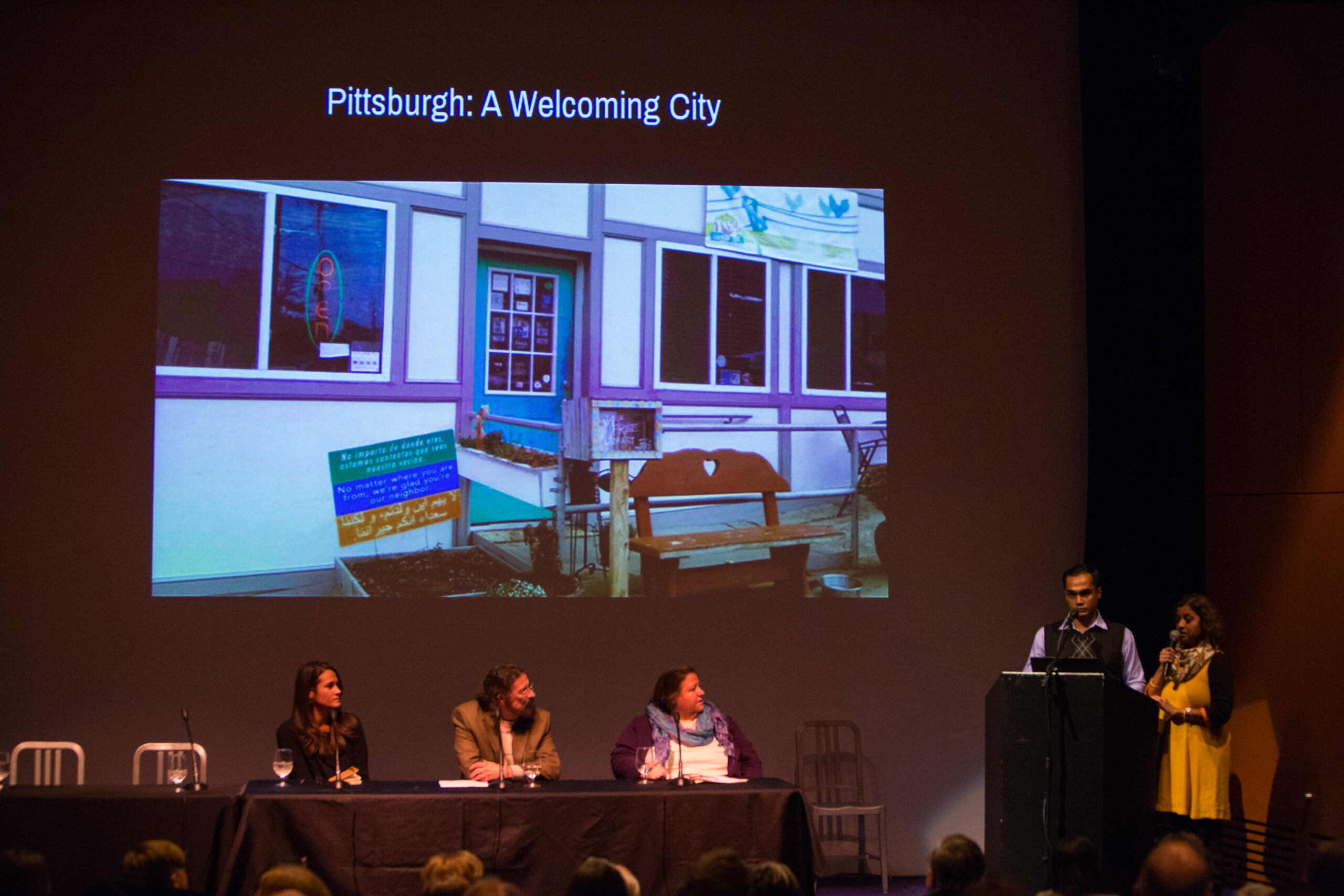 Three panelists sit at a table on the left side of a stage. They look towards two speakers standing behind a lectern to the right. Projected behind them is a photo of a storefront with a green, blue, and yellow sign in front. Above the image is the text Pittsburgh: A Welcoming City.