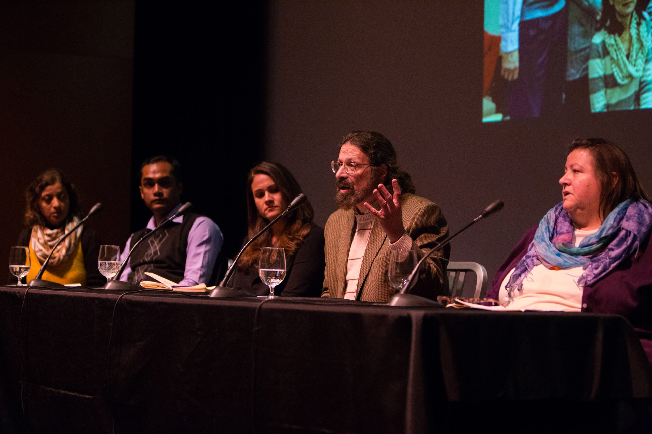 Five panelists sit at a table on stage. One of them is speaking and gesticulating as the others look on.