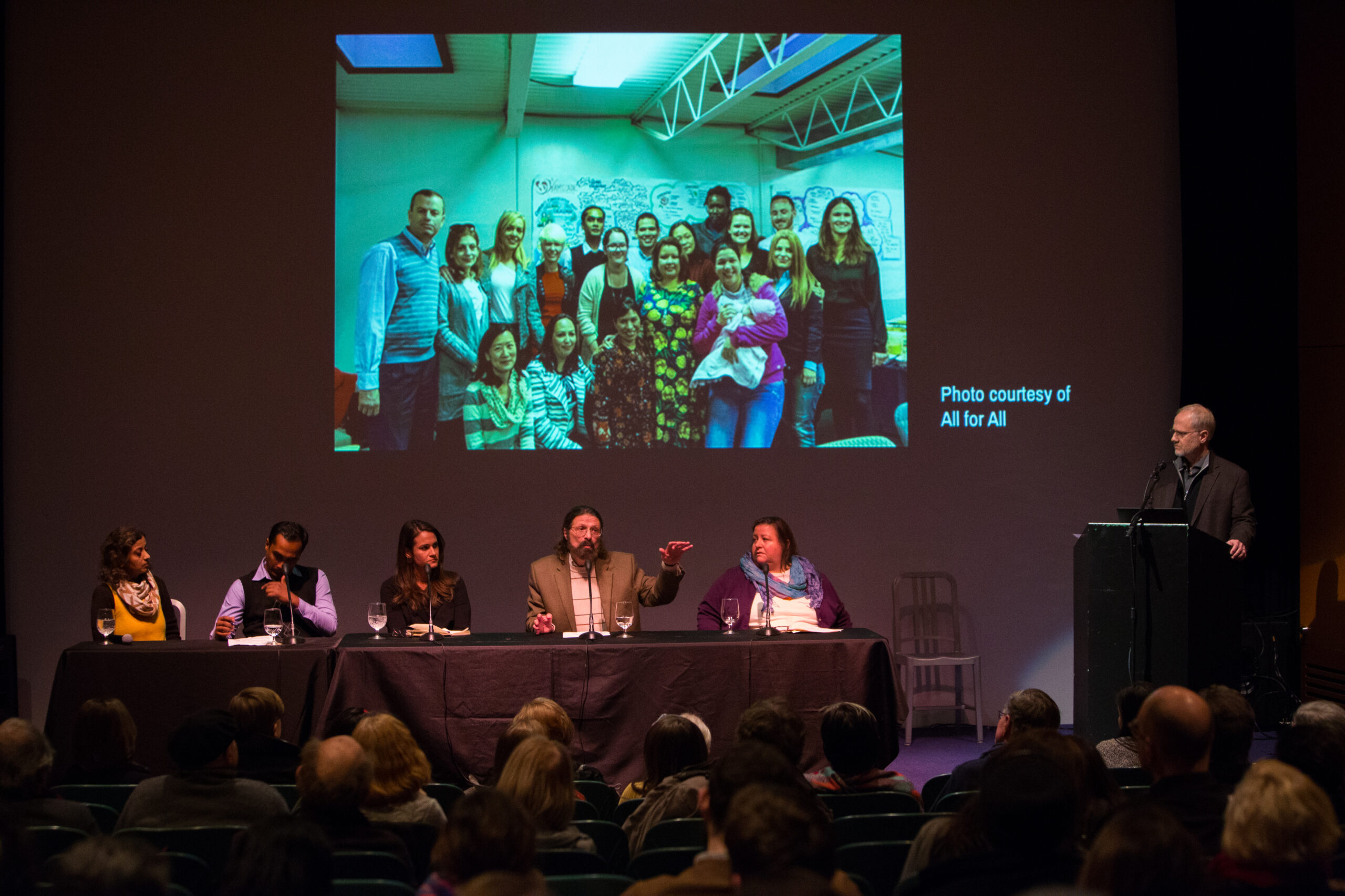 Five panelists sit at a table on the left side of a stage, and one man stands behind a lectern on the right side. One man at the table is talking and gesticulating, as the others look on. Behind them is projected a photo of a group of people posing for the camera. To the side of the image is the text Photo courtesy of All for All
