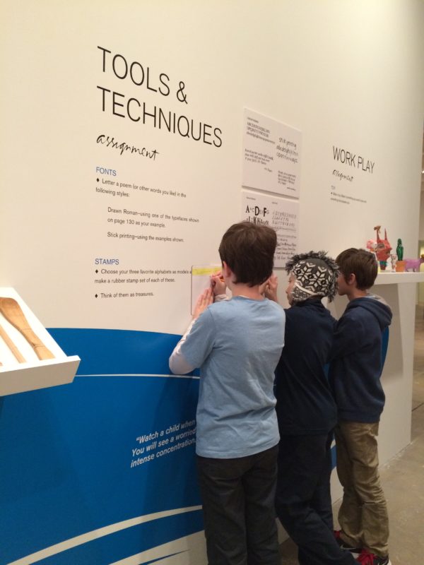 Image of 3 students writing in journals, leaning against a wall. The wall text behind them reads “tools and techniques” and “work and play”.