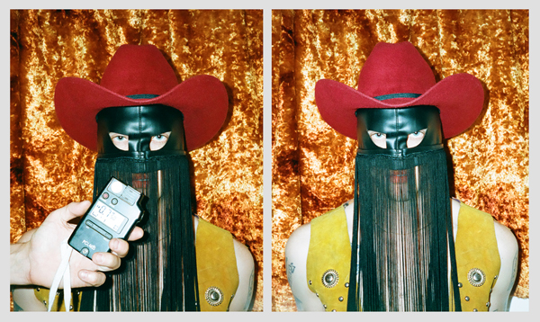 A double image of a man wearing a red cowboy hat, yellow vest, and a green mask with fringe hanging from it stands in front of a brown backdrop. In the image on the left, there is a hand holding a light meter in front of the man.