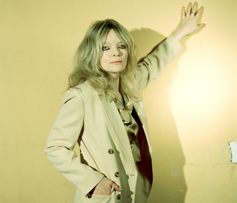 A woman with long, blonde hair stands in front of a yellow wall with her left hand on the wall above her head. She is wearing a coat and her right hand is in the front pocket of the coat.