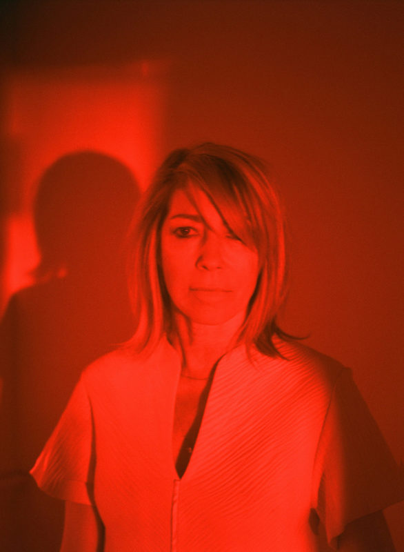 A photo of a woman with blond hair looking towards the camera. The photo is in a red tint.