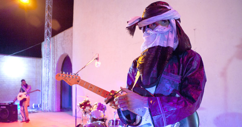 A man standing in front of a white wall holds a guitar. There is another man with a guitar in the background as well as a drum set.