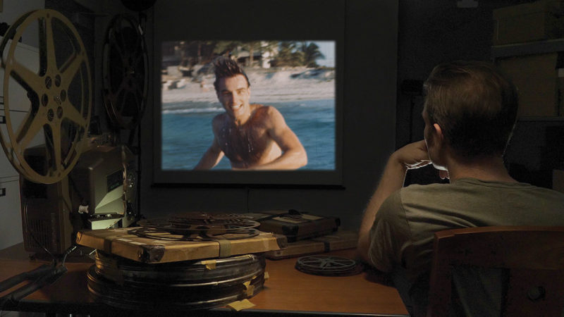 Footage of Montgomery Clift smiling at the beach is projected on the wall in the background while co-director Robert Clift watches the footage in the foreground. On a table next to Robert are stacks of film reels and an old film projector.