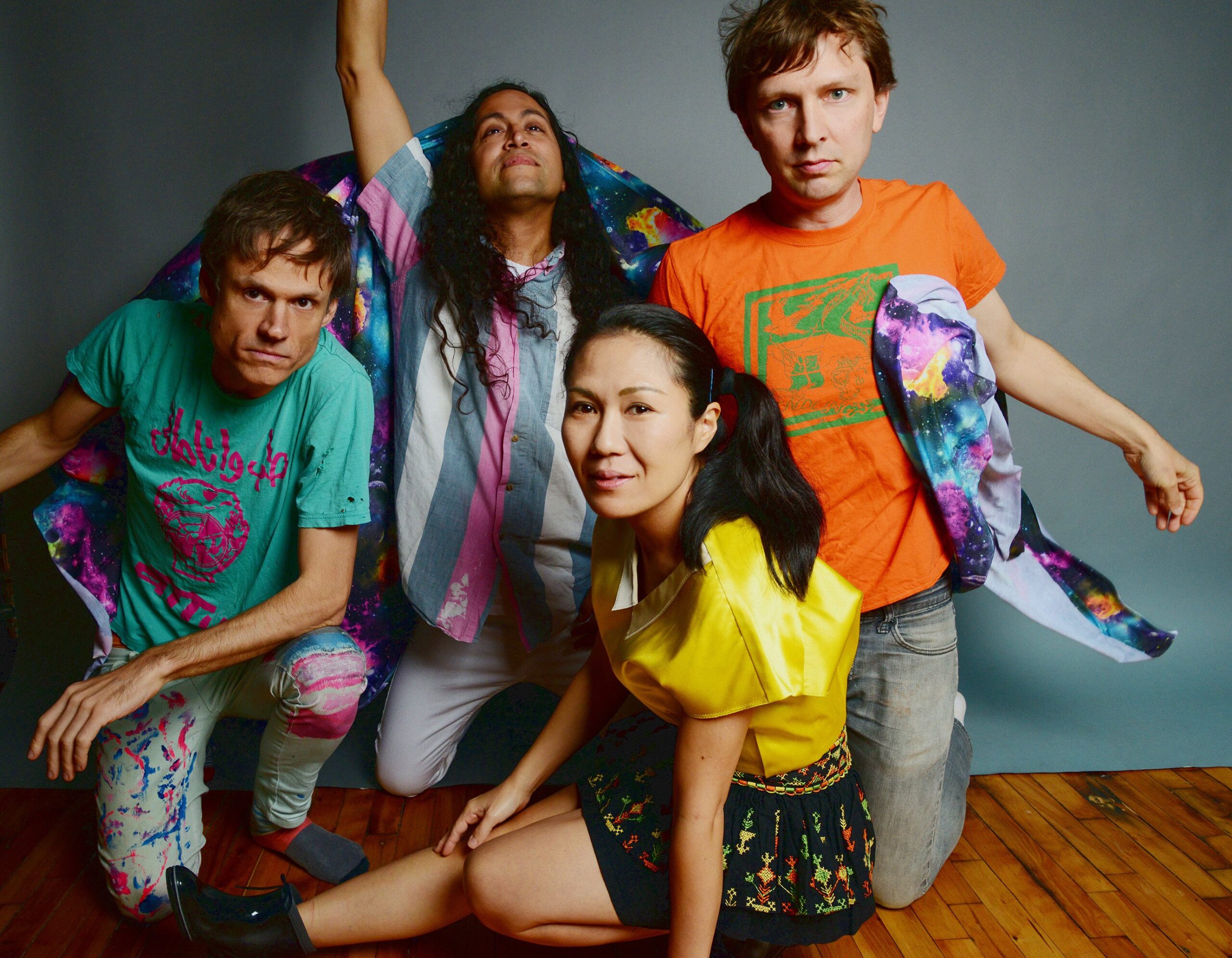 A group of people in various colorful outfits sit or kneel on a hardwood floor in front of a gray background.