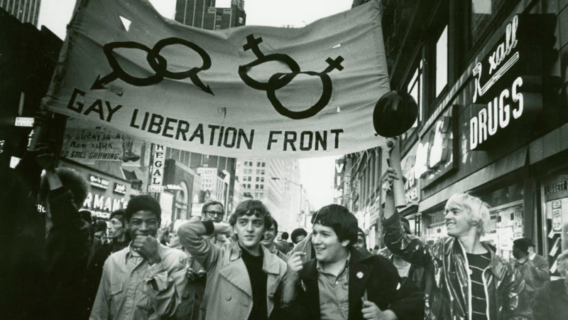 A black and white film still of people marching down a city street below a banner that says, Gay Liberation Front.