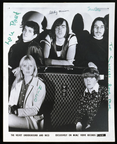 Black-and-white photograph on white coated paper. It shows the members of the Velvet Underground and Nico: Lou Reed, Sterling Morrison, John Cale, Nico, and Moe Tucker, posing around a Vox brand speaker. It is signed in green felt-tip and black ink: The Velvet Underground.