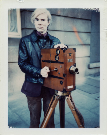 A color Polaroid photograph of Andy Warhol, standing outside next to a large, wooden, movie camera on a tripod. He is wearing a black leather jacket over what appears to be a purple shirt, and brown pants.