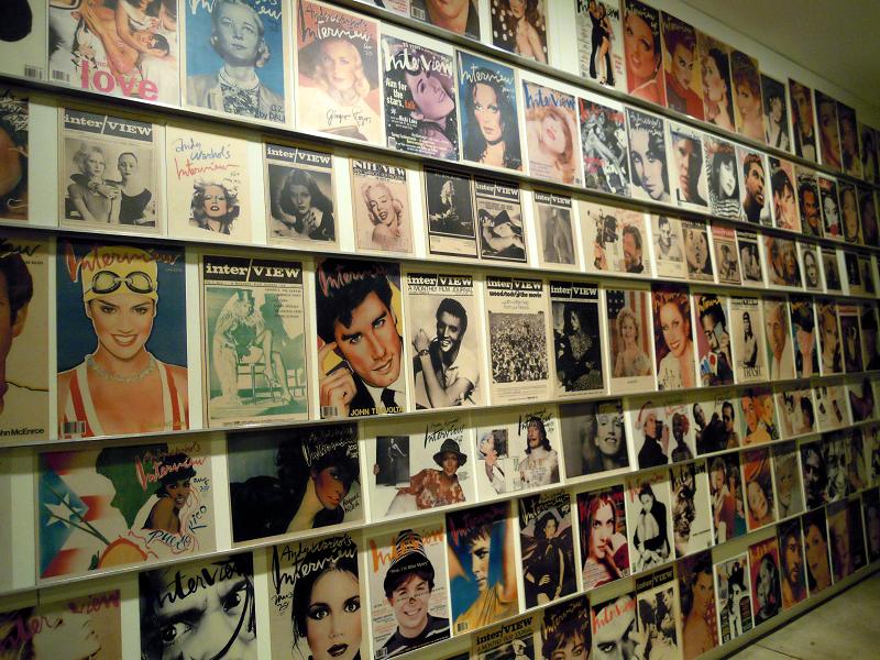 A color photo of an Interview magazine installation at The Andy Warhol Museum. There are multiple magazine covers displayed on seven rows of shelving.