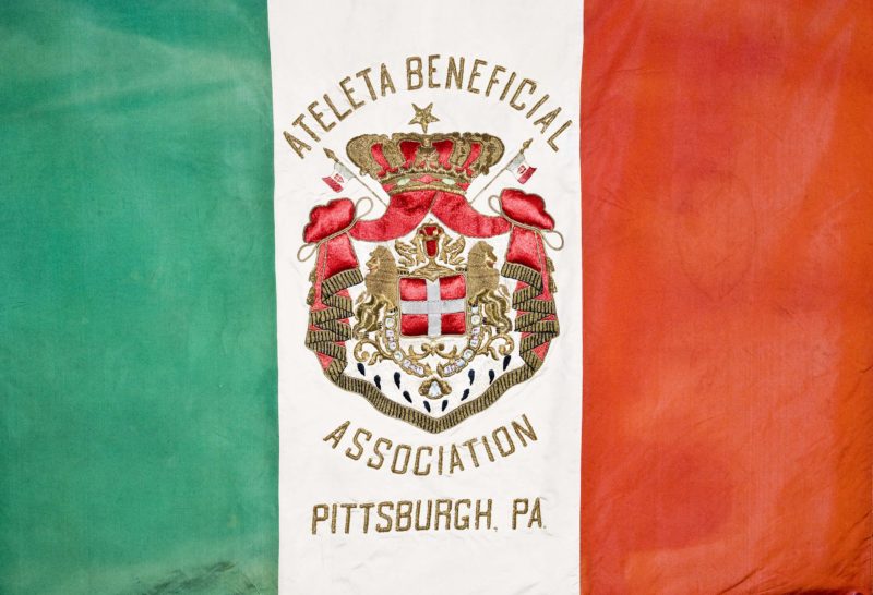 Front view of an Italian flag, with green, white, and red vertical stripes from left to right. Within the white strip, there is a red and gold seal with the word “Ateleta Beneficial Association, Pittsburgh, PA.” stitched in.