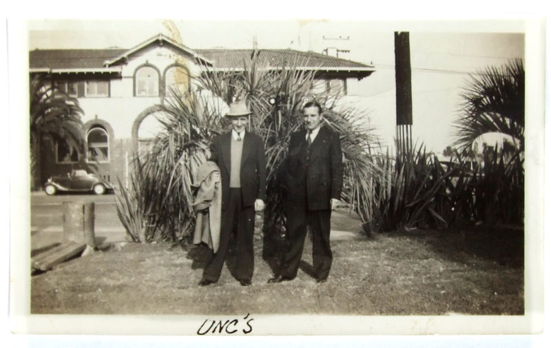 Sepia photograph of two men in suits standing in front of plants in a yard. A two-story home behind the men, across the street, faces the camera.