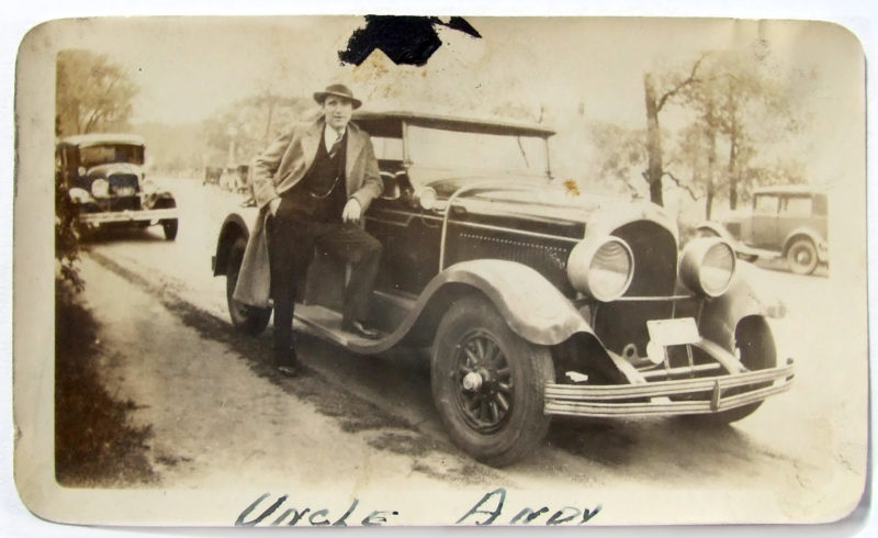 Sepia photograph of a man in a suit and trench coat, posing with one foot on a car.