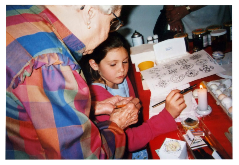 A woman instructs a young girl on Pysanky techniques. The woman is wearing a multi-colored blouse.