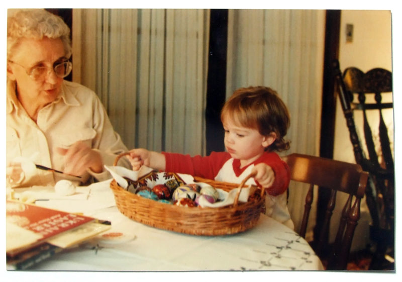 A woman is in the process of painting an egg, while a toddler looks at a basket full of completed pysanky eggs.
