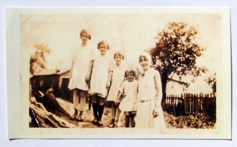 Sepia photograph of five young girls in light dresses smiling at the camera.