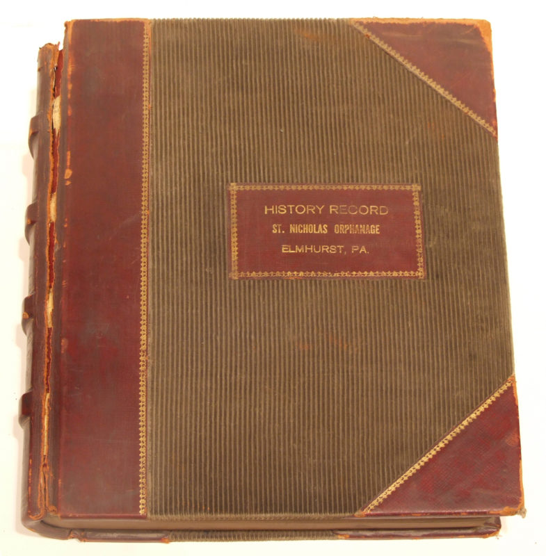Front view of History Record of the St. Nicholas Orphanage. The Record is a leather bound book that is red and brown.