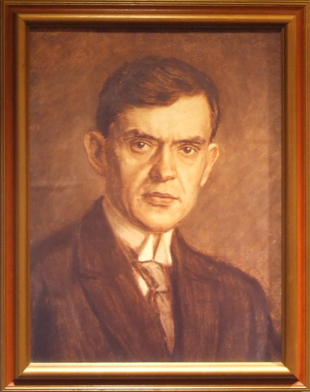 Framed painted portrait of a man with short brown hair and brown eyes. He is wearing a black suit with a gray tie and white shirt.