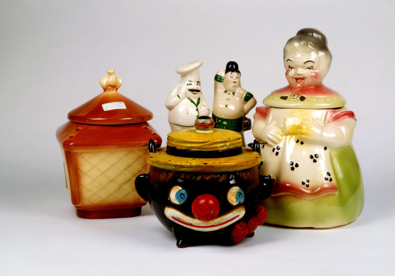 A collection of cookie jars. The cookie jar on the right is in the shape of a plump woman wearing a green dress with her hair in a bun. The jar in the middle is a clown wearing a hat with a bulbous red nose, red bowtie, and blue eyes. The jar on the left is in the shape of a lantern with a white bird on top.