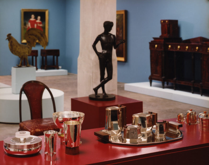 Silver cups, creamers, coffee pots, and bowls sitting on a wooden table in a gallery. Behind the table is a black statue of a man standing as well as a large statue of a rooster. Two paintings hang on the wall beside a large wooden armoire.