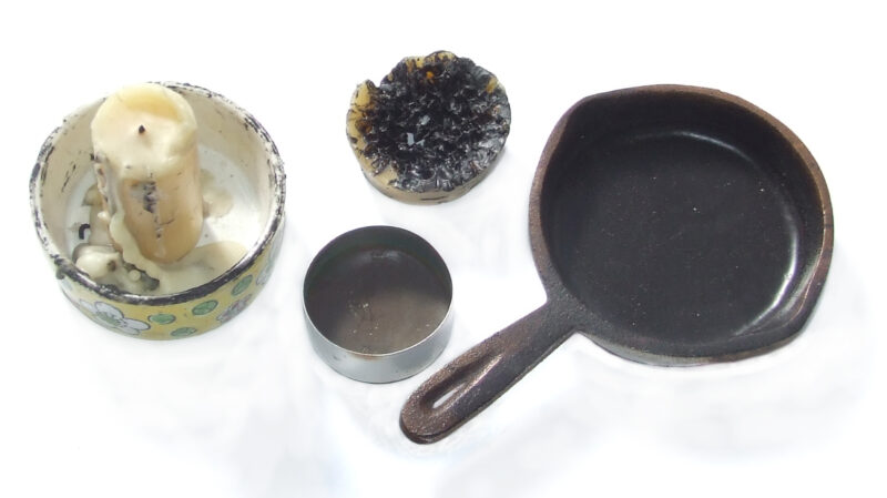 Photo of tools used for wax melting. Tools pictured are a miniature cast iron pan, a small silver container, and a partially used candle.