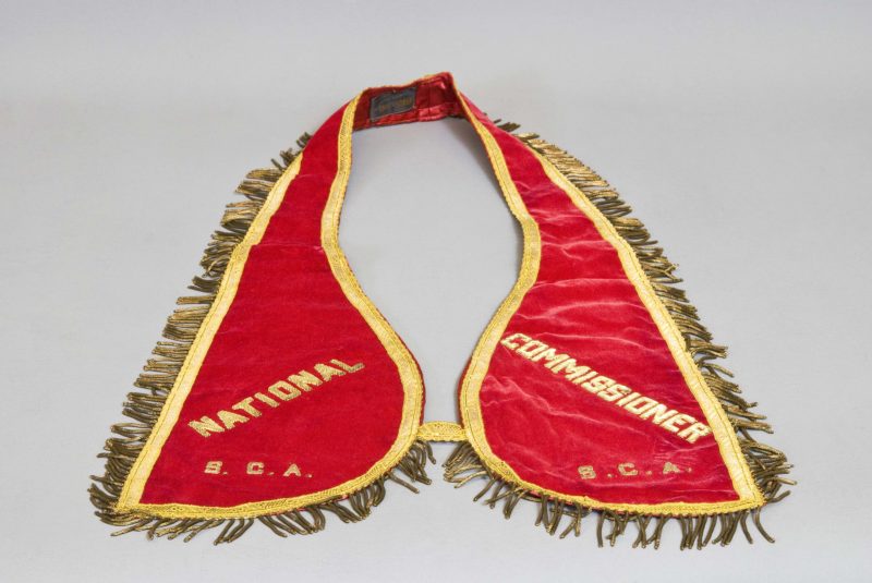 Front view of red sash with gold piping and fringe. The sash reads “National Commissioner” in gold letters.