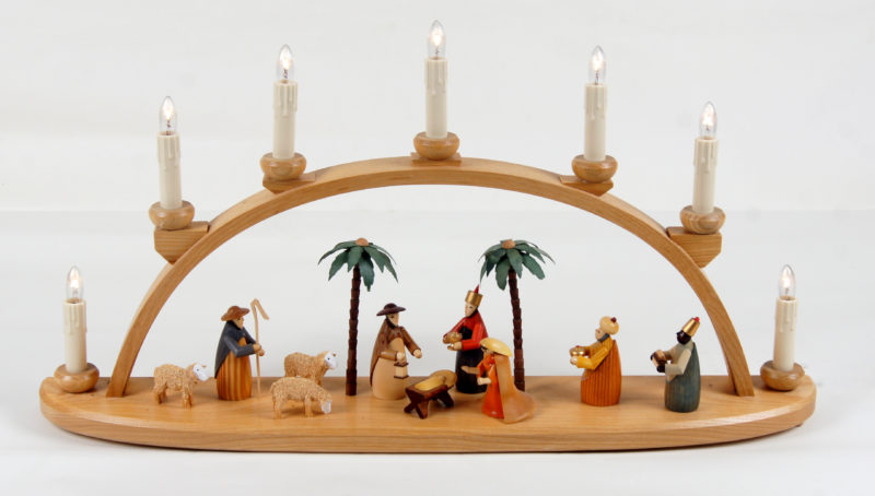 Image of candle arch with seven candles. The arch arcs over figurines of various Christmas figures, sheep, and palm trees.