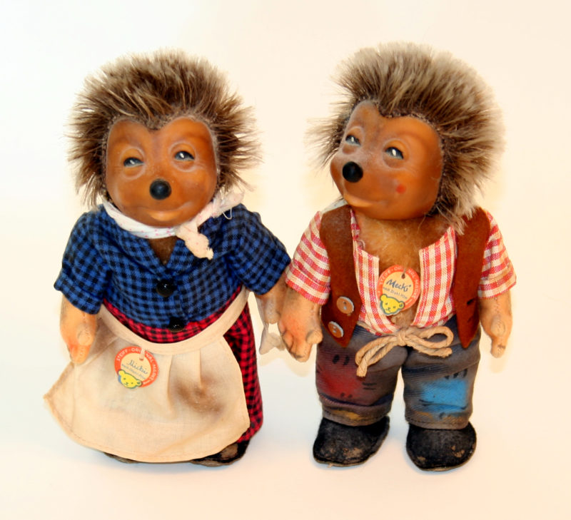Two hedgehog figurines stand upright in human clothing, next to one another. The female figurine wears a checkered scarf, blouse, and pants along with an apron while the male figurine wears an open shirt and vest.