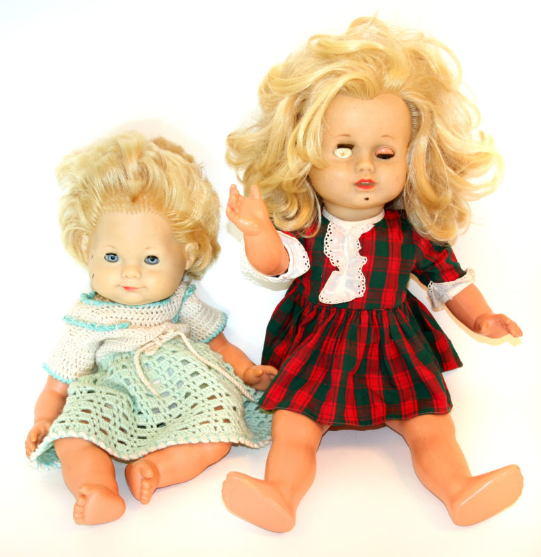 Front image of two blonde baby dolls sitting next to each other. The doll on the left has blue eyes and a crocheted dress, and the doll on the right is wearing a dress and has an eye that’s been worn off.