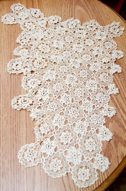 Above view of a small baptism blanket placed on a wooden table. The blanket is crocheted, white, and features a floral pattern.