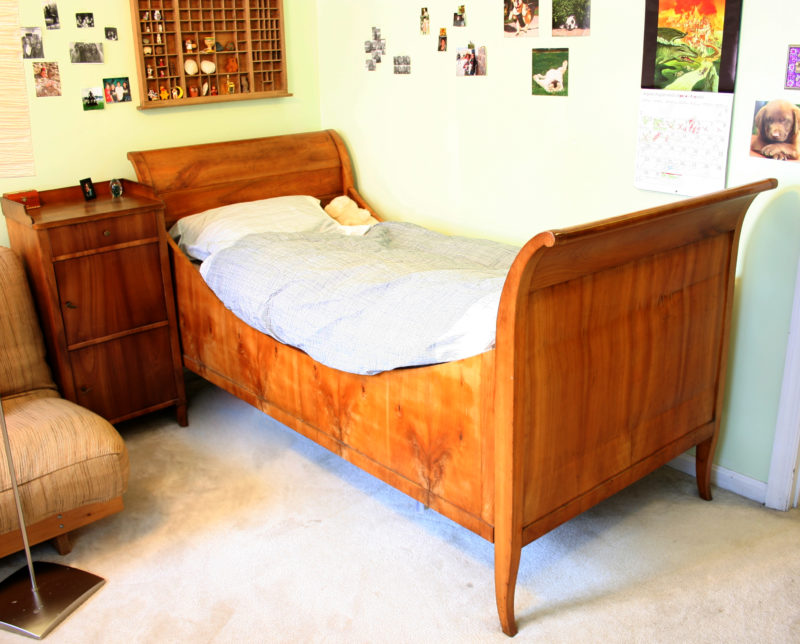 A Biedermeier style wooden bed sits in a corner of a child’s bedroom, nestled between a wall and a nightstand. On the bed is a white pillow and blue comforter; photos, a calendar, and trinkets cover the walls.