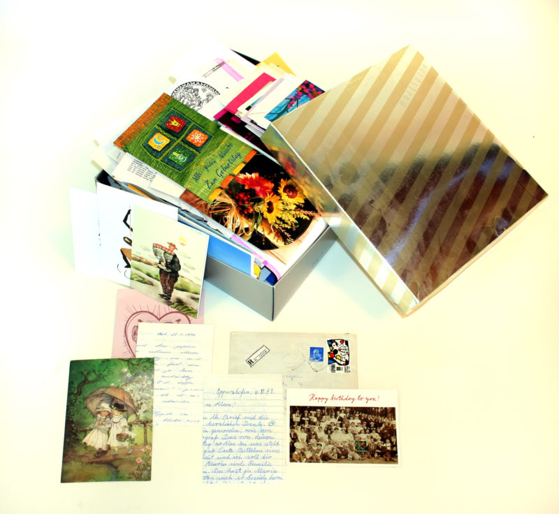 A box of letters is displayed with full selected letters and postcard images visible. The box’s removable gold lid is visible and leans on the full box of letters.