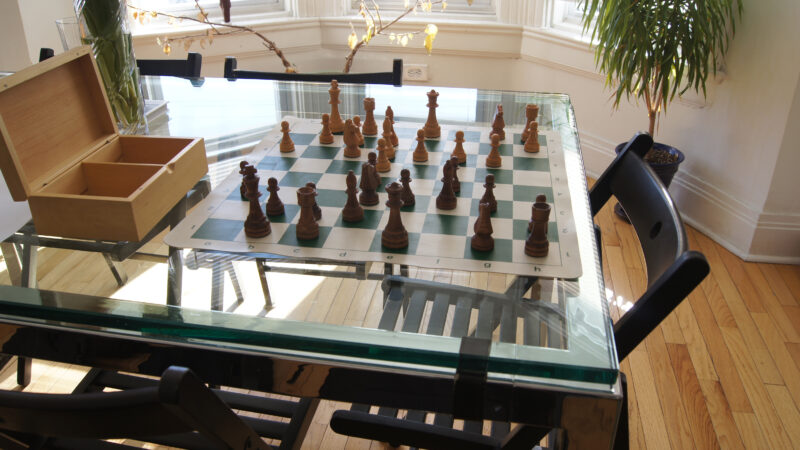 Image of chess set on display on a glass table inside a home. The chess pieces are wooden and sit atop a foldable, portable chess board next to a wooden box.