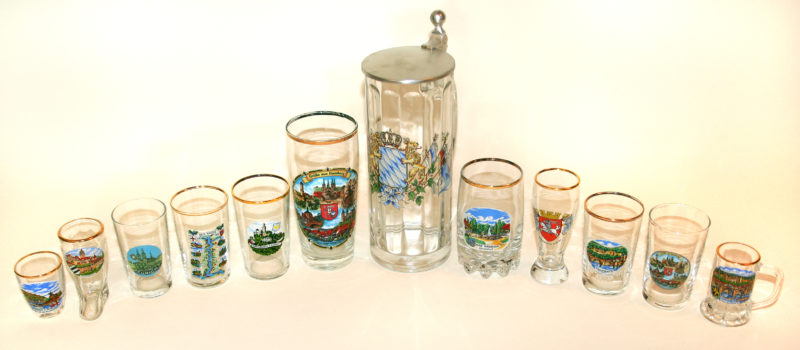 Front view of twelve collectible jars arranged by size, with the two largest glasses in the middle and glasses getting smaller from the middle, outward. The glasses have decorative rims and images of illustrated landscapes on their fronts.