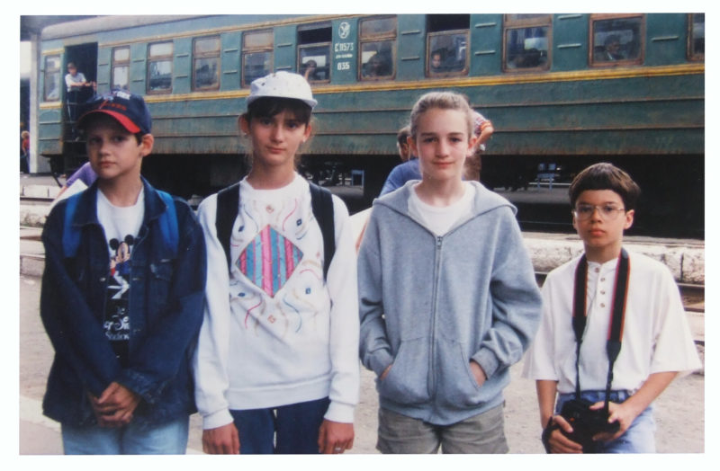 Photograph of four children standing near a green train and facing the camera.