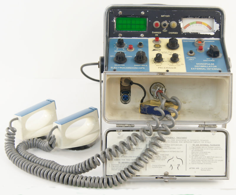 Front view of a 1960s defibrillator. The defibrillator’s monitor is multicolored and the defibrillator paddles extend out of the machine, connected by thick, gray, spring-shaped cables.