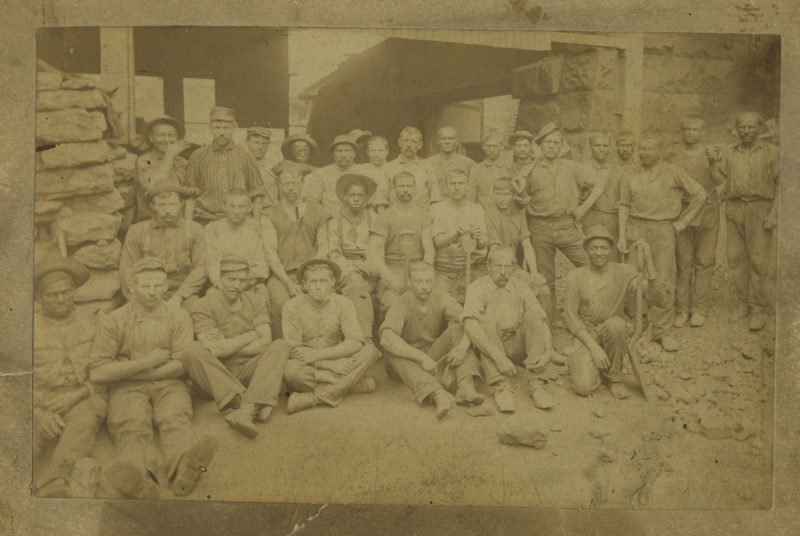Sepia photograph of crewmembers at Jones & Laughlin Steel Company. There are approximately thirty men, both white and African-American, seated or standing in rows. Some men are holding shovels.
