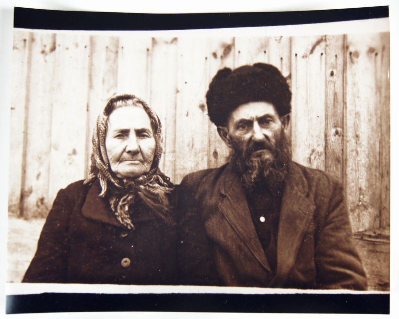 iew of sepia photograph of a senior woman and man seated next to one another. The woman is wearing a scarf around her head and the man is wearing a fur hat.