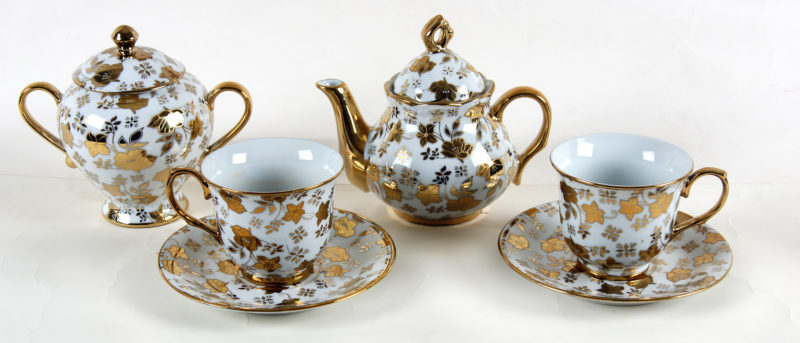 Front image of a tea set with ornate, gold-on-white floral patterns. The set includes a teapot, two tea cups and dishes, and a spoutless pot.
