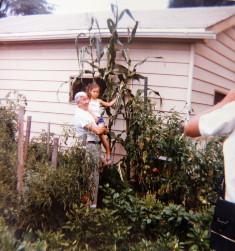 Photo of a senior man holding up a young girl, posed inside of a garden full of plants and high stalks. The garden grows outside of a cream-colored house.