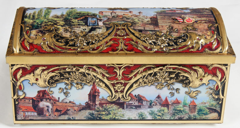 Front view of a closed tin box with ornate gold embellishment and a painted village.