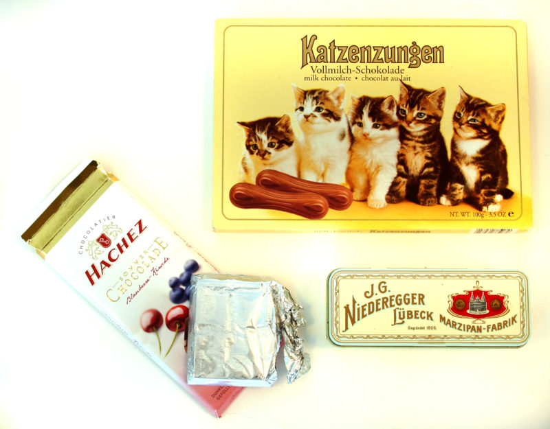 Front view of three packages of chocolate. One box of chocolates has an image of five kittens on the front, another is a small tin box, and the third is a wrapped bar of chocolate with a partially unwrapped silver package of chocolate on top.