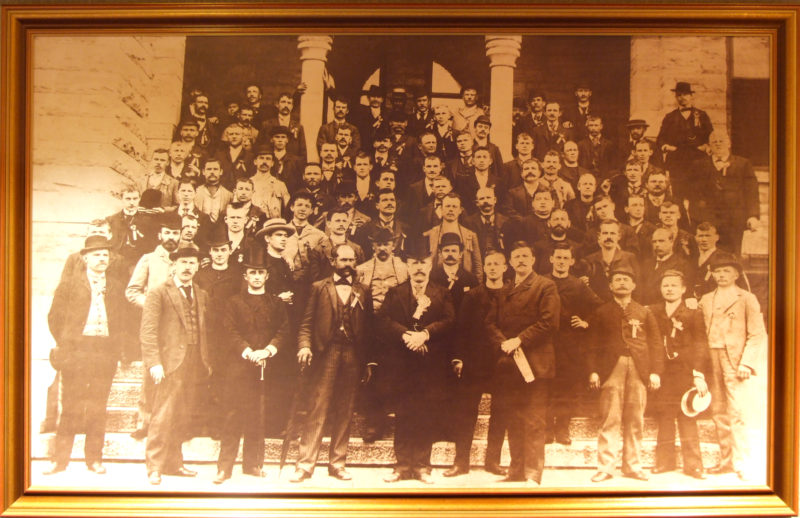 Framed sepia photograph of a large group of men lined up on steps outside of a building with two columns.