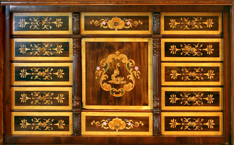Close up-view of the ornate design that backs a wooden desk. The design is made up of eleven rectangular boxes that border a square box in the backing’s center.