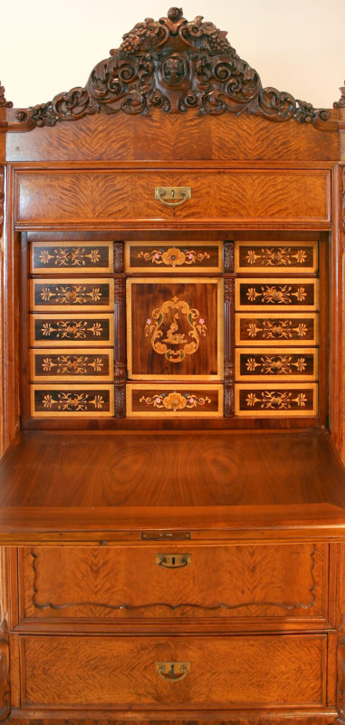 Front view of opened heirloom desk. The largest compartment has been opened, revealing a cubby with an ornate design with gold, pink, and green accents backing the negative space of the desk.