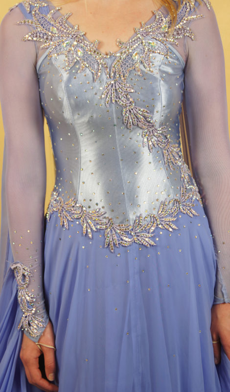 View of sequinned details of a blue dress’ torso. The material of the dress’ torso is metallic and fitted—sequins are spread across the torso and an ornate embellishment lines and sashes the dress’ torso.