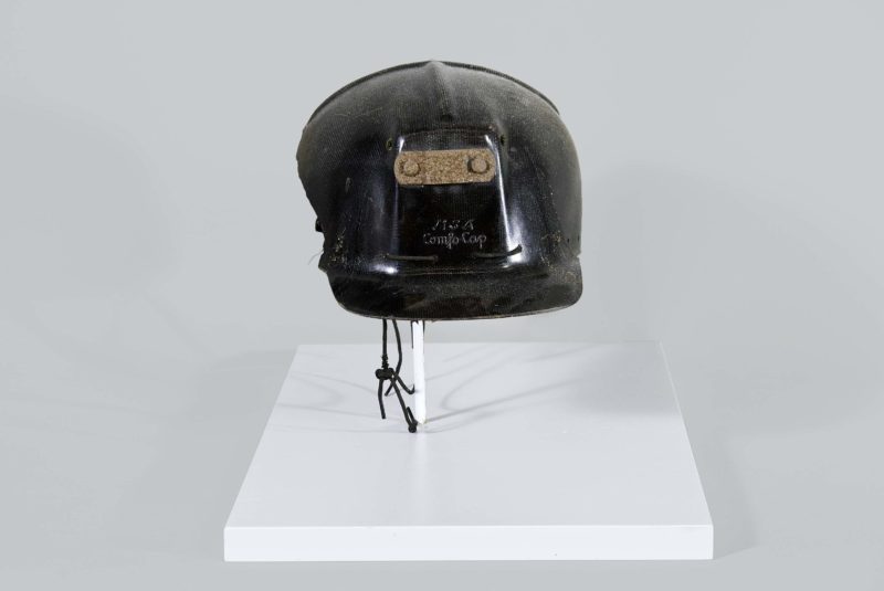 Side view of metal miner’s helmet. The helmet is black and shaped like a baseball cap, and the helmet’s side has been broken.